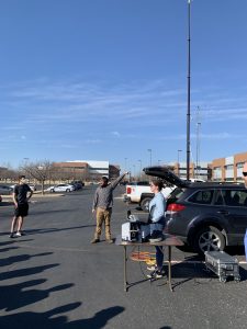 Wesley T. Honeycutt, the author, points at a tall instrument mast next to a Subaru in a parking lot. His colleague Elizabeth Spicer stands nearby. They are teaching meteorology instrumentation with pieces of equipment they use in their research. A student looks on.