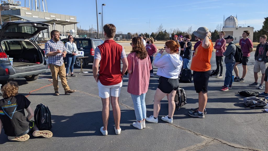 A class of students look on as Wes instructs them about instrument theory while his colleague Elizabeth Spicer looks on in the parking lot outside the National Weather Center in Norman, OK. They are teaching meteorology instrumentation for the OU course.
